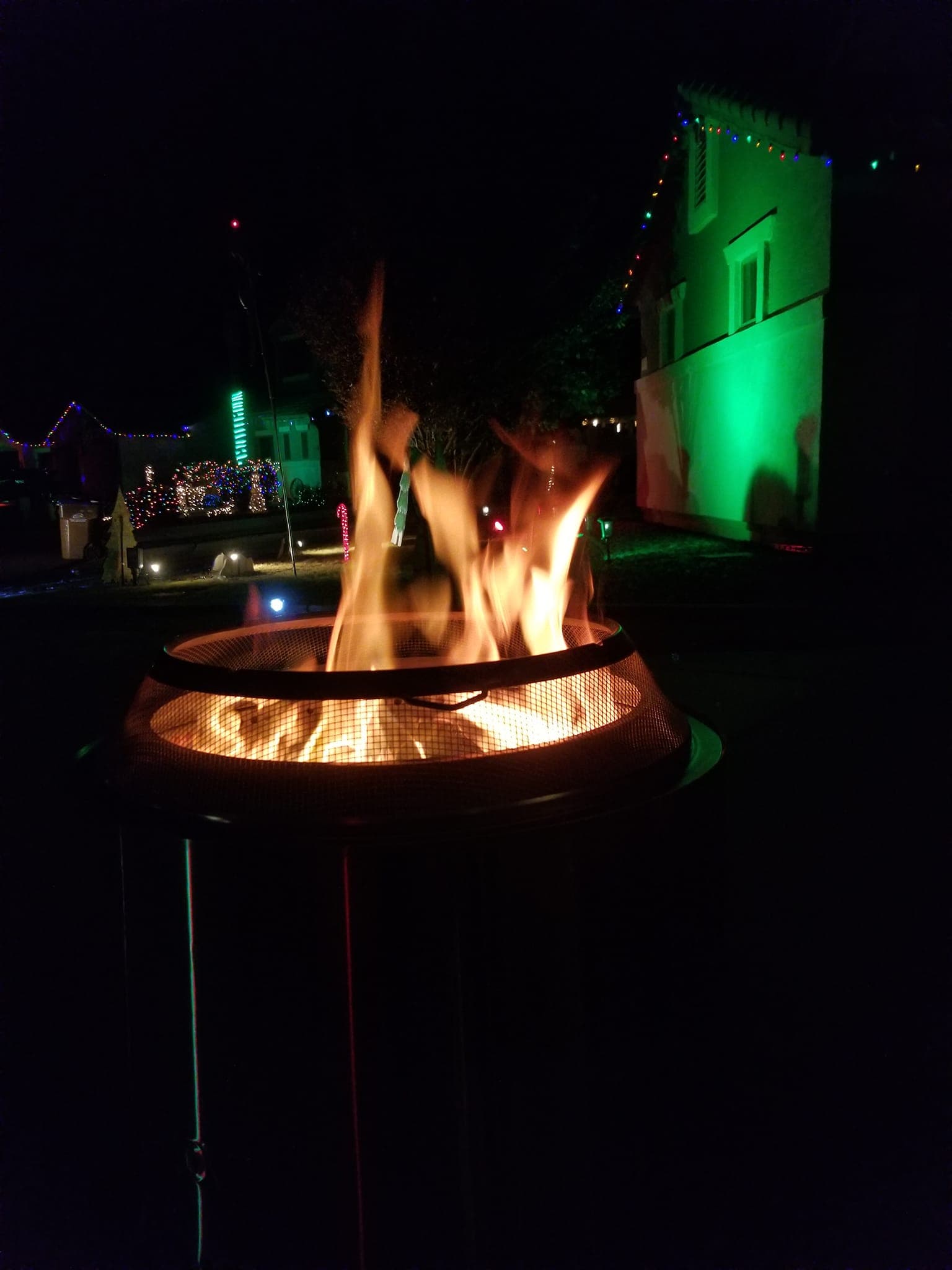 A fire pit glowing in front of green-washed homes and holiday lights
